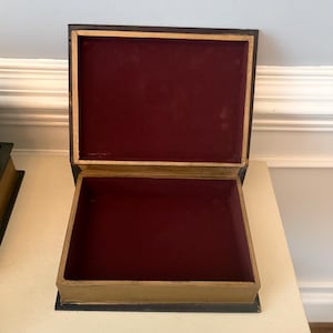 May include: An open, rectangular box with a hinged lid. The exterior is black with gold trim, and the interior is lined with a deep red velvet material. The box is sitting on a white surface.