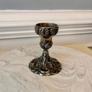 May include: A silver candlestick with a detailed grape and vine design. The candlestick has a wide base, a slender stem, and a cup-shaped top. The metal has a tarnished appearance, adding to its vintage charm. The candlestick is likely made of silver or a silver-plated metal.