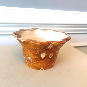 May include: A small, handcrafted ceramic pot with a scalloped rim. The exterior is a warm brown with speckled orange and white accents. The interior is a creamy white. A decorative piece for plants or small items.