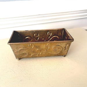 May include: A rectangular brass planter with a tapered shape and four small ball feet. The exterior surface features an embossed scrolling vine and leaf pattern. The metal has a weathered, aged patina with visible dark spots and texture.