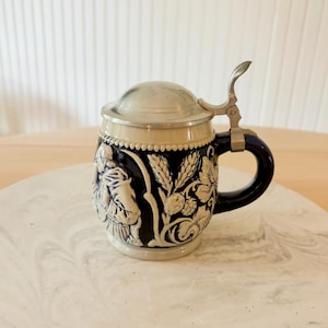 May include: A dark blue ceramic beer stein featuring a metallic lid and handle. The exterior displays a raised, cream-colored relief depicting a figure and botanical elements including wheat stalks. The stein has a smooth, rounded base and top.