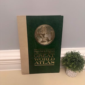 May include: A vintage Reader's Digest Great World Atlas. The book has a green cover with a gold embossed world map and text. The spine is a light beige color. A small potted plant sits to the right of the book.