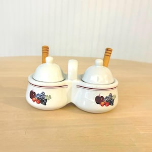 May include: A white ceramic double condiment server with two matching lids and two wooden spoons. The base features a thin maroon horizontal stripe and a colorful graphic print of red apples and purple grapes on each side of the container.
