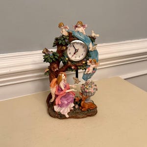 May include: A decorative table clock featuring a round white face with black numerals and hands. The clock is integrated into a resin sculpture depicting a tree with green leaves, three small cherubs, white doves, a seated angel, and a deer.