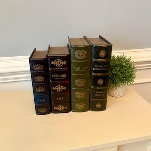 May include: Four decorative books with gold lettering and accents. The books are in shades of brown and green, with titles including "Sister Carrie" and "War and Peace." A small potted plant sits to the right of the books on a cream-colored surface.