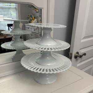 May include: A white, three-tiered cake stand with a scalloped edge design. The stand has a pedestal base and is made of ceramic. The stand is displayed on a white surface, with a mirror reflecting the stand in the background.
