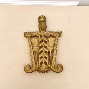 May include: Ornate gold-colored metal trivet with a decorative design. The trivet features a central vertical element with a series of leaf-like shapes, flanked by two stylized torch-like elements. The design includes a loop at the top for hanging and curved flourishes at the bottom.
