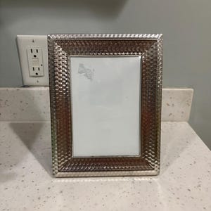 May include: A rectangular picture frame with a metallic, hammered texture finish. The frame features a wide, reflective border with a repeating dimpled pattern surrounding a central white rectangular insert with a small gray mark.