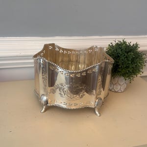 May include: A silver-toned metal bowl with a scalloped top edge featuring a pierced lattice design. The exterior displays an engraved crest and floral motifs, supported by four small, curved feet with a beaded trim along the base and rim.