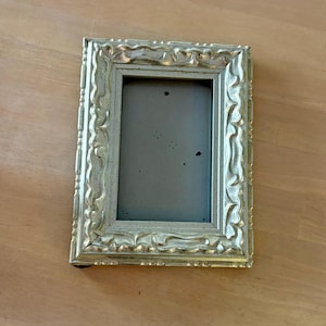 May include: A rectangular picture frame with a metallic silver finish and a raised, ornate, scalloped border design. The frame features a smooth, light gray backing insert with several small, dark, scattered spots visible on the surface.
