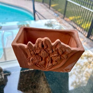 May include: A square terracotta planter with a floral relief design on the front. The planter is a warm reddish-brown color and has a slightly rough texture. The floral design is detailed and adds a decorative element to the planter.