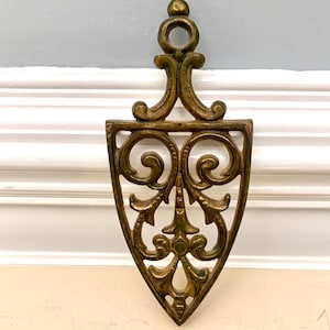 May include: Ornate, vintage-style trivet in a gold tone. The trivet features a shield shape with intricate scrollwork and a loop at the top for hanging. A decorative kitchen accessory.