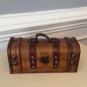 May include: A small, wooden treasure chest with a curved top and a dark brown leather handle. The chest is adorned with dark brown leather straps and brass-colored hardware. The front features a decorative clasp.