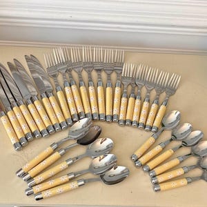 May include: A flatware set consisting of stainless steel knives, forks, and spoons. Each piece features a pale yellow handle decorated with a repeating white daisy pattern. The set includes multiple knives, forks, and spoons arranged in groups.