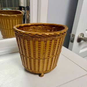 May include: A woven wicker basket with a rounded top and a flared base. The basket is light brown and has a textured surface. The basket has three small feet. The basket is sitting on a white surface.