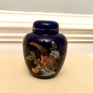 May include: A navy blue ceramic jar with a lid, featuring a gold rim. The jar is decorated with a detailed illustration of a pheasant, flowers, and foliage in shades of gold, orange, and green. The jar has a glossy finish.
