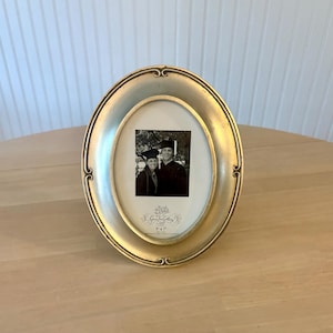 May include: An oval-shaped gold picture frame with decorative scroll accents at the top and bottom. The frame contains a black and white photograph of two graduates and a label that reads Green Tree Gallery and 5 x 7 inches at the bottom.