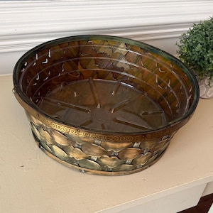 May include: An oval-shaped decorative basket with a bronze-colored metal trim and a leaf-like pattern. The basket has a clear, brown-tinted interior and is designed with a decorative, ornate edge. A small potted plant is visible in the background.