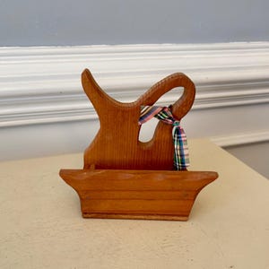 May include: Wooden decorative piece with a basket-like base and a curved handle. A colorful plaid ribbon is tied around the handle. The wood has a warm, natural finish, and the piece sits on a light-colored surface.