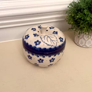 May include: A ceramic apple-shaped lidded jar with a cream-colored base and a hand-painted pattern of small blue flowers and green leaves. The lid features a sculpted stem and a single leaf design, with a deep blue band encircling the center.