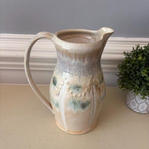 May include: A ceramic pitcher with a curved handle and a pouring spout. The exterior features a glazed finish in shades of cream, soft gray, and muted green, with vertical raised ridges and small dot accents encircling the midsection of the body.