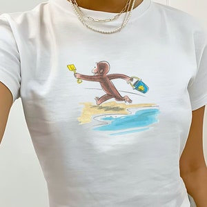 May include: White t-shirt with a graphic of a monkey running on the beach with a bucket and shovel. The monkey is brown and the bucket is blue.