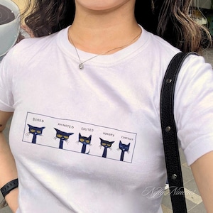 May include: White t-shirt with a graphic of five black cats with different facial expressions. The text above each cat reads "Bored", "Annoyed", "Excited", "Hungry", and "Content".