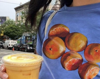 May include: A blue t-shirt with a graphic print of orange slices. The person is holding a clear plastic cup with orange juice.
