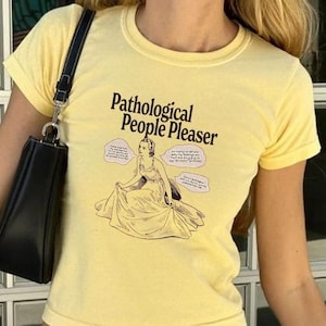Könnte beinhalten: Ein gelbes T-Shirt mit kurzen Ärmeln mit dem Text "Pathological People Pleaser" und einer Cartoon-Illustration einer Frau in einem Kleid. Das Shirt wird mit einer hellblauen gestreiften Hose kombiniert.