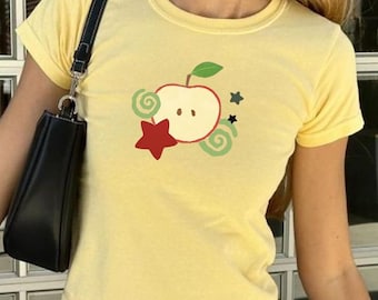 May include: A yellow t-shirt with an apple design, worn with light blue and white striped trousers. A black handbag is carried on the shoulder. The person is holding a drink in a clear cup.