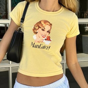 Puede incluir: Una camiseta amarilla recortada con una ilustración vintage de una mujer y el texto "ManEater!" impreso en la parte delantera. La mujer lleva un vestido rojo y sostiene una taza con un diablo rojo dentro. La camiseta se combina con pantalones de rayas azules claros.