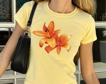 Puede incluir: Una camiseta amarilla recortada con estampado floral naranja. La camiseta se combina con pantalones de rayas azules.