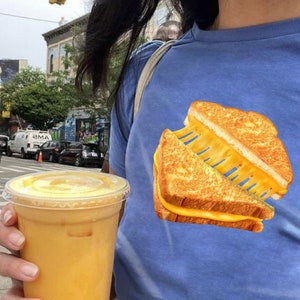 以下が含まれることがあります： 青いTシャツを着て、グリルチーズサンドイッチのグラフィックが描かれた人。その人は、透明なプラスチックカップに入った黄色の飲み物を手に持っている。