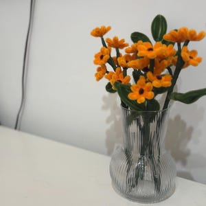 Op de afbeelding: Een heldere glazen vaas met een getextureerd oppervlak bevat een boeket van oranje viltbloemen met groene stelen.