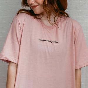 May include: A light pink t-shirt with the text "professional yapper" in black. The shirt has a crew neck and short sleeves. The model is wearing a brown hat.