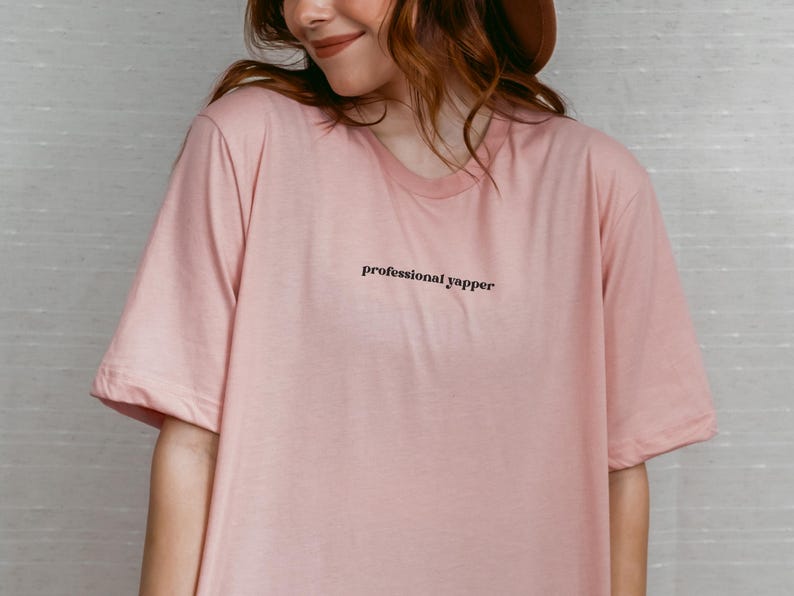 May include: A light pink t-shirt with the text "professional yapper" in black. The shirt has a crew neck and short sleeves. The model is wearing a brown hat.