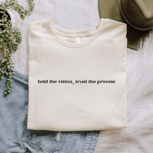Op de afbeelding: Een crèmekleurig T-shirt met korte mouwen en de zwarte tekst "hold the vision, trust the process" in het midden op de borst. Het shirt heeft een ronde hals en ligt plat neergelegd met een gevouwen rand aan de onderzijde.