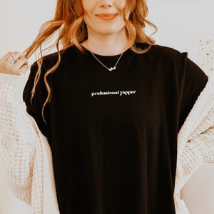 May include: Black t-shirt with the text "professional yapper" in white. The shirt is worn with a white cardigan and a delicate necklace. The model is smiling and holding their hair.