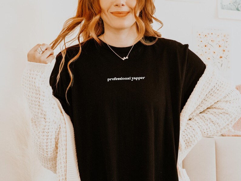 May include: Black t-shirt with the text "professional yapper" in white. The shirt is worn with a white cardigan and a delicate necklace. The model is smiling and holding their hair.