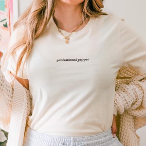 May include: Cream-colored t-shirt with the text "professional yapper" in a black, minimalist font. The shirt is paired with a cream-colored cardigan and light gray sweatpants. A gold necklace is visible.