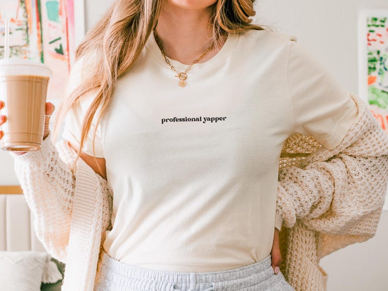 May include: Cream-colored t-shirt with the text "professional yapper" in a black, minimalist font. The shirt is paired with a cream-colored cardigan and light gray sweatpants. A gold necklace is visible.