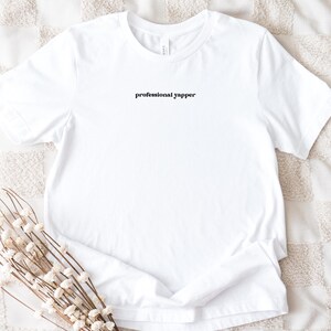 May include: A white t-shirt with the text "professional yapper" in a black, stylized font. The shirt is displayed on a textured, off-white surface, with a sprig of dried flowers in the foreground.