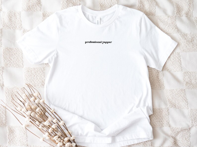 May include: A white t-shirt with the text "professional yapper" in a black, stylized font. The shirt is displayed on a textured, off-white surface, with a sprig of dried flowers in the foreground.