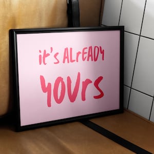 Könnte beinhalten: Ein gerahmter rosa Druck mit den Worten "it's ALREADY YOURS" in roter Schrift. Der schwarze Rahmen kontrastiert mit dem rosa Hintergrund. Der Druck wird auf einer braunen Oberfläche angezeigt.