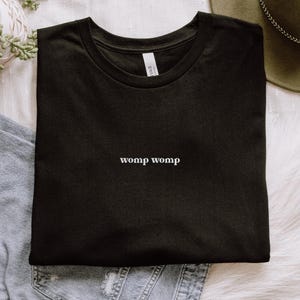 Puede incluir: Camiseta negra de cuello redondo con la palabra "womp womp" en blanco, centrada en el pecho. La camiseta está doblada sobre una prenda vaquera. Un sombrero verde y una planta son visibles en el fondo.