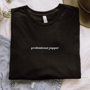 May include: Black t-shirt with the phrase "professional yapper" in white, centered on the chest. The shirt is folded neatly, and the background includes a plant, a hat, and denim.