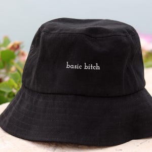 Puede incluir: Sombrero de pescador negro con el texto bordado "basic bitch" en blanco. El sombrero está hecho de una tela suave y tiene un ala ancha. El fondo presenta una vista borrosa de agua y flores rosas.