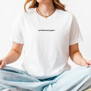 May include: White t-shirt with the text "professional yapper" in a black, simple font. The model is wearing light blue jeans and a gold necklace. The person is sitting cross-legged.