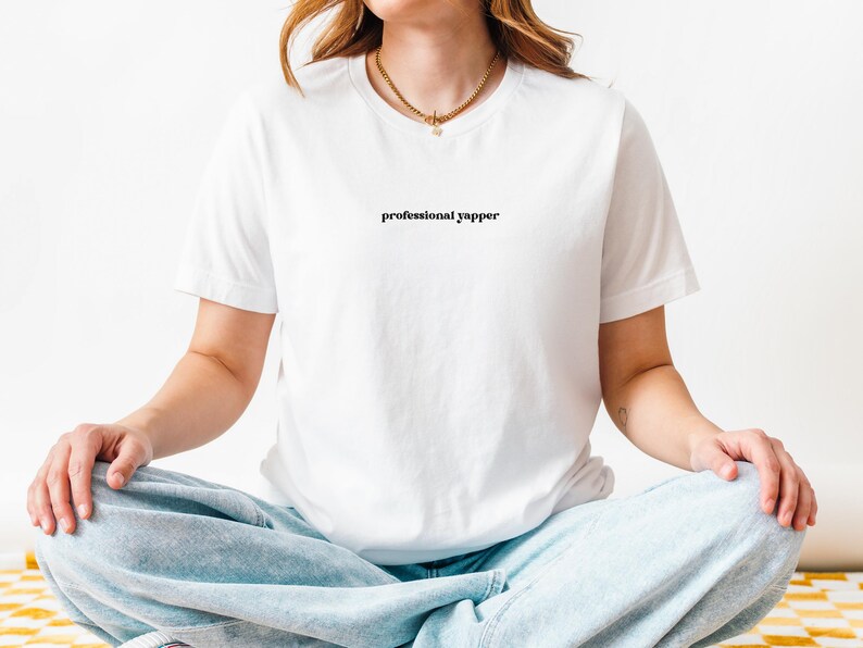 May include: White t-shirt with the text "professional yapper" in a black, simple font. The model is wearing light blue jeans and a gold necklace. The person is sitting cross-legged.