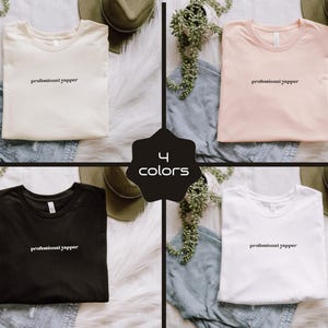 May include: Four t-shirts in cream, pink, black, and white, each with the text "professional yapper" in a simple font. The shirts are displayed on a white fur-like surface with denim and hats.